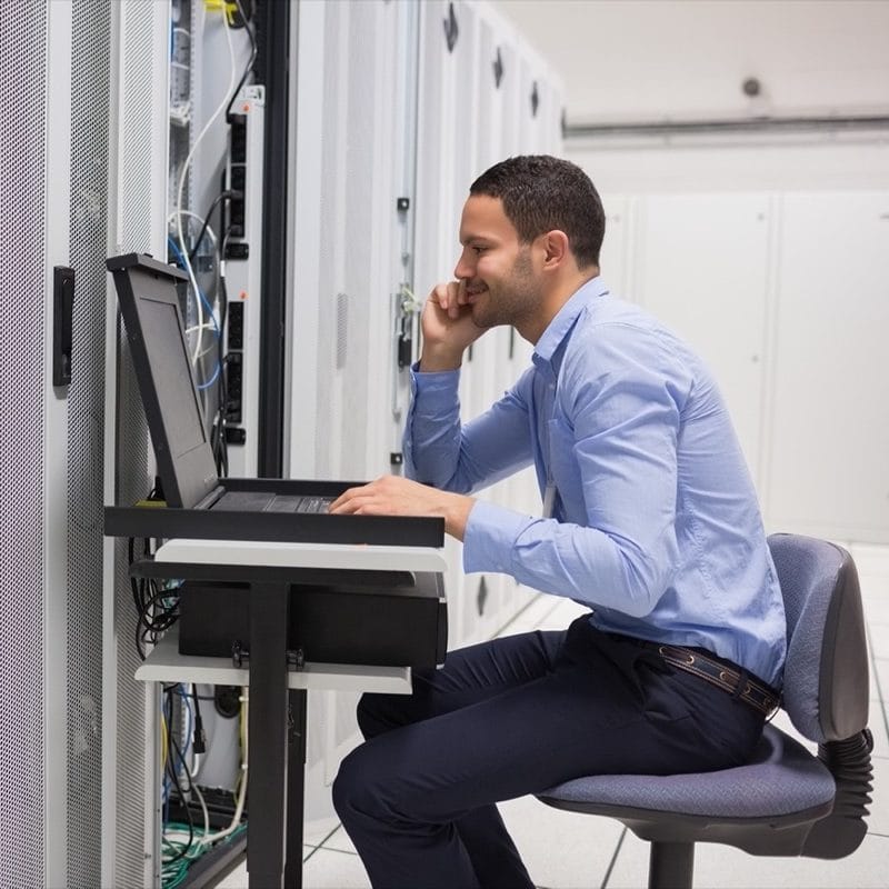 IT professional sitting and working a laptop computer in a server room.