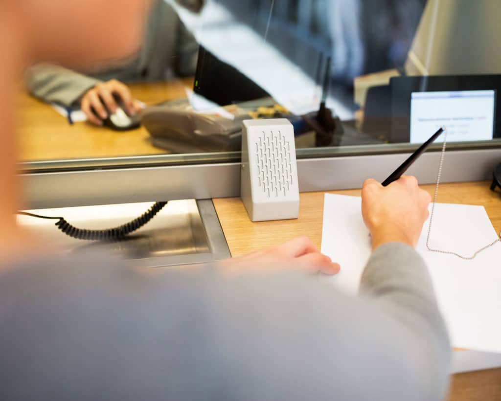 Image of a bank teller desk. On one side they are filling out paperwork. On the other side of the desk the person appears to be working on their computer.