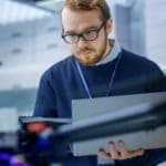 Engineer wearing glasses and blue sweater golding laptop computer while looking at drone.