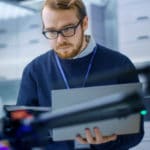 Engineer wearing glasses and blue sweater golding laptop computer while looking at drone.