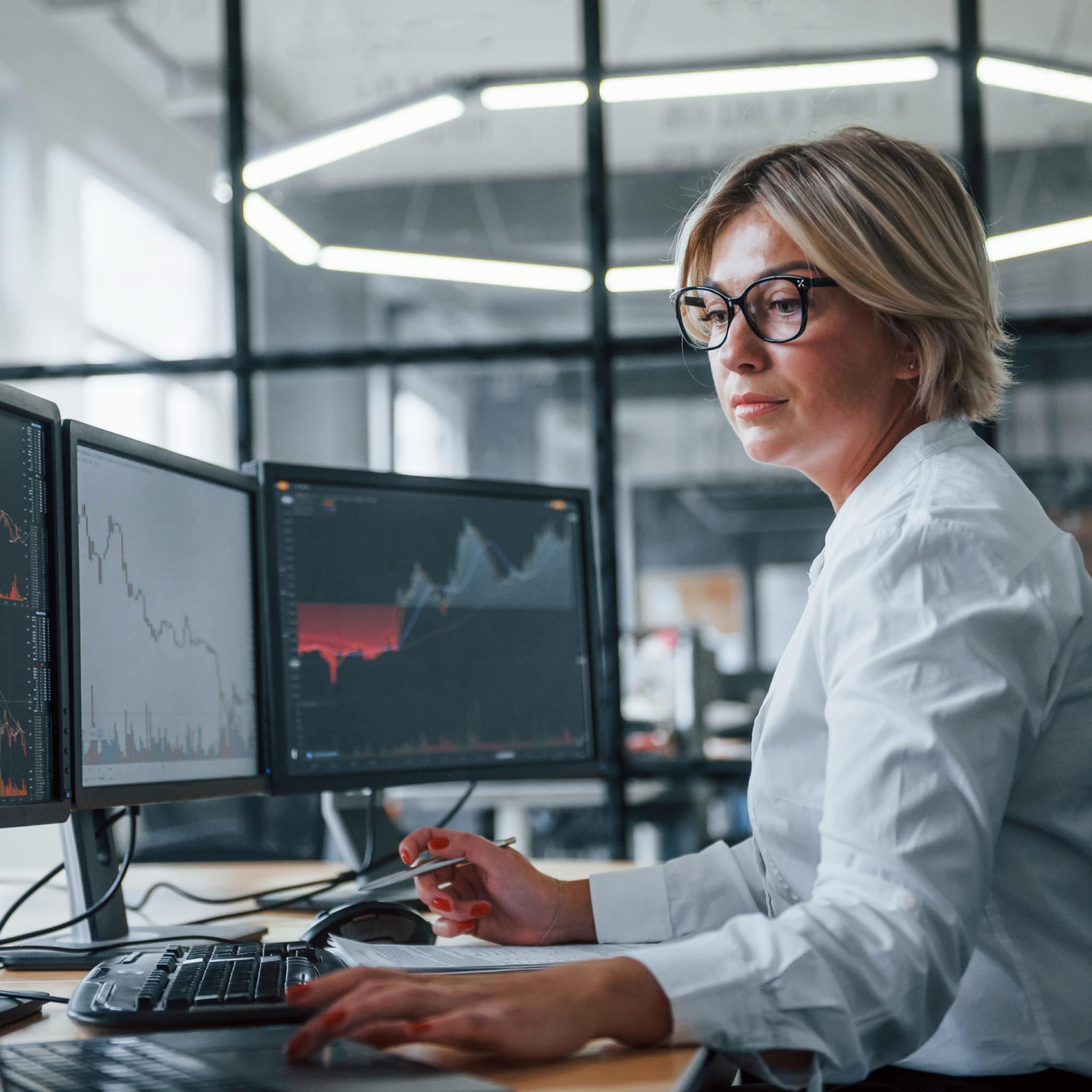 Female stockbroker wearing glasses working on laptop computer beside monitors displaying graphs.