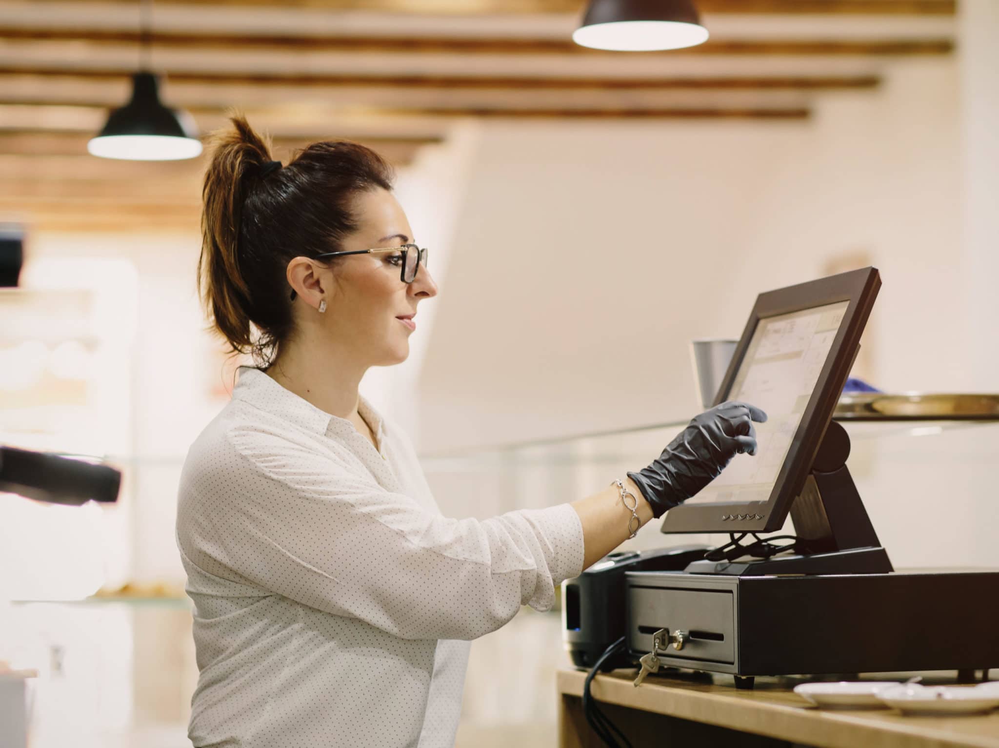 Woman wearing glasses pressing buttons on POS machine in cafeteria.