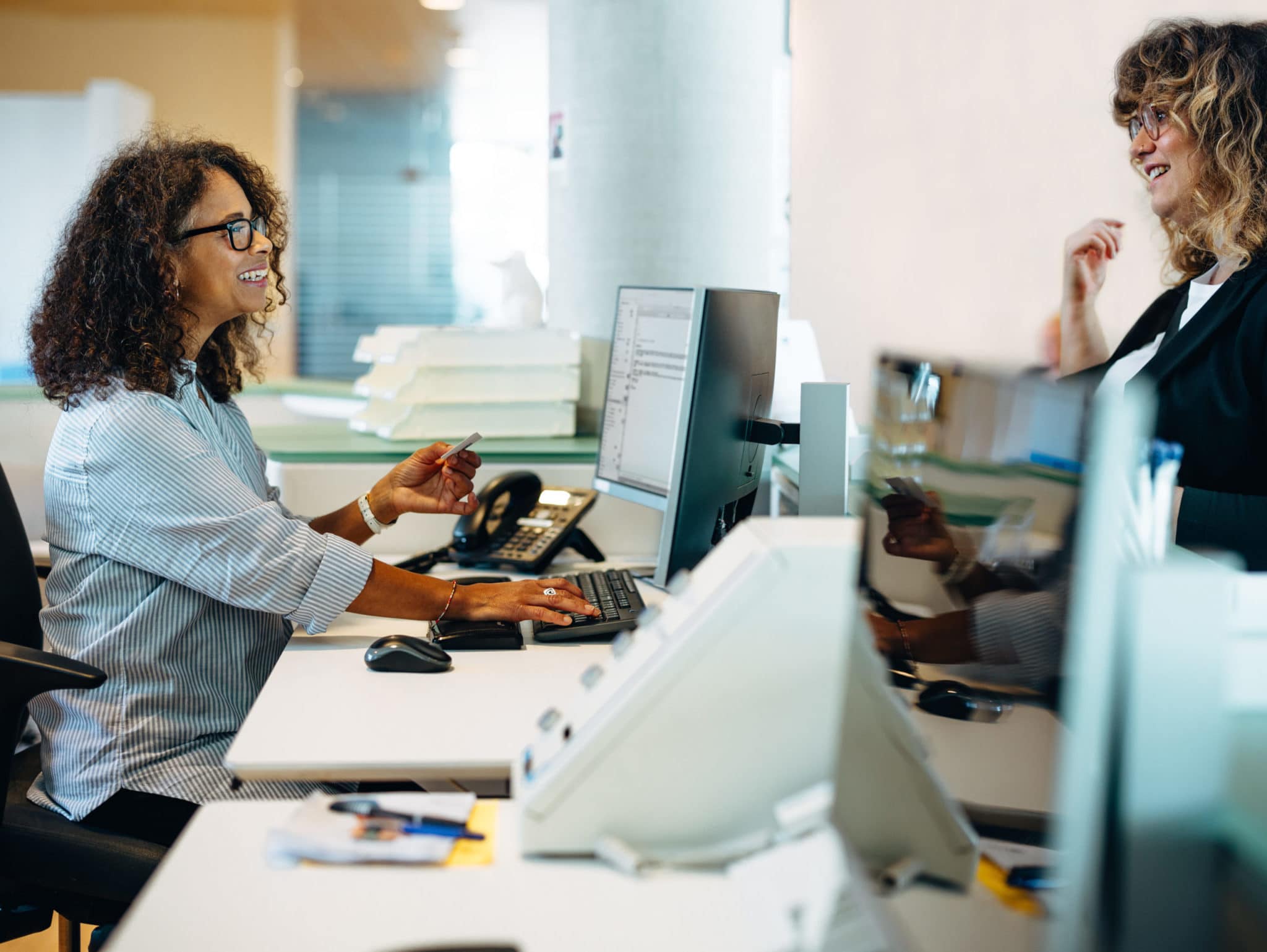 Woman office administrator sitting at desk and smiling while assisting a woman standing at her desk.