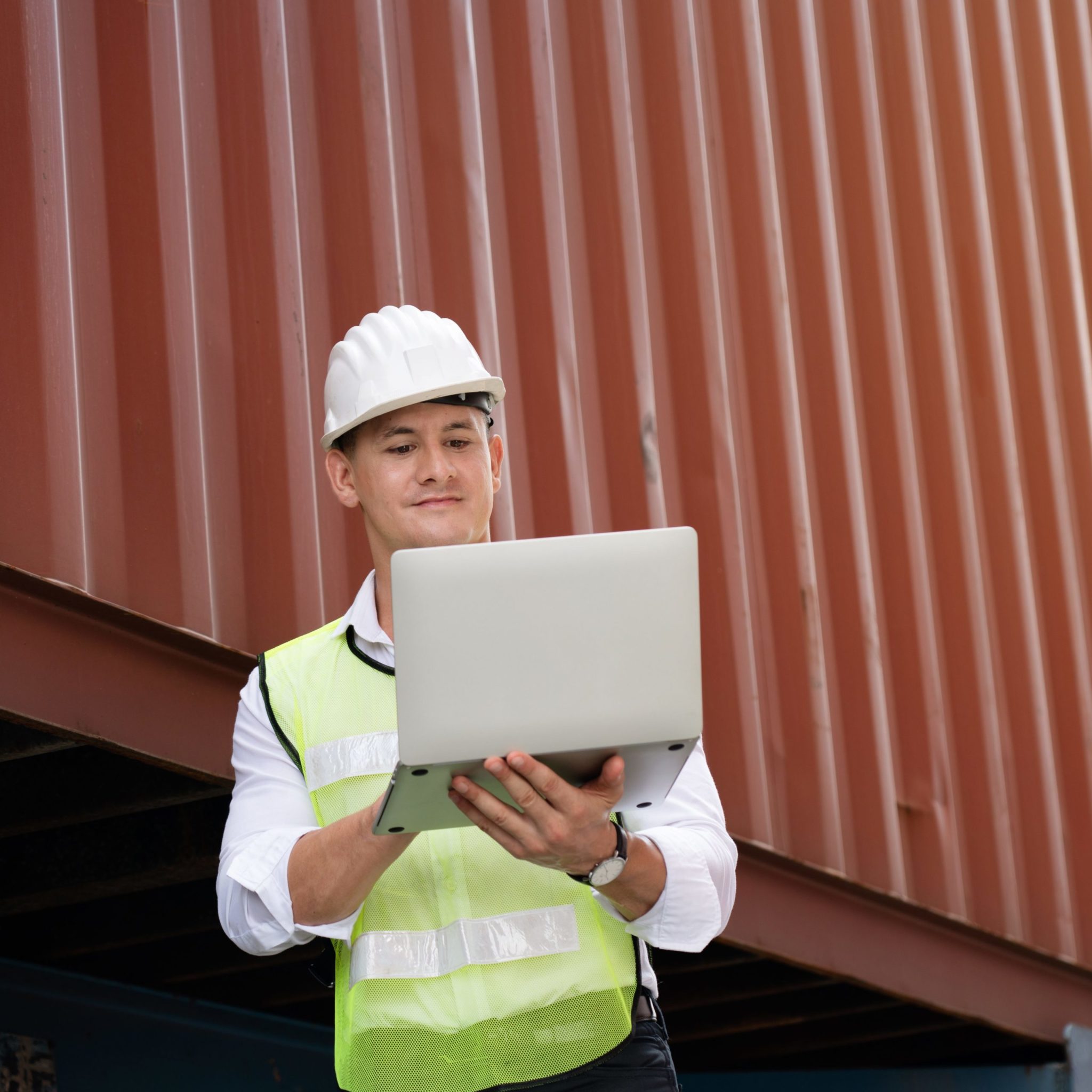 Male engineer in hardhat and reflective vest holding laptop computer and working on it.