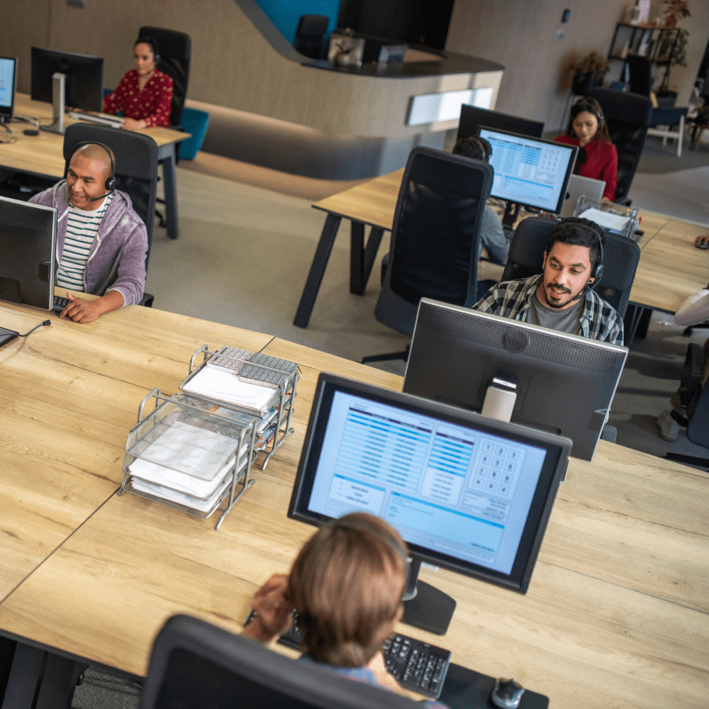 Office setting with everyone wearing headsets while seating at desks working on computers.