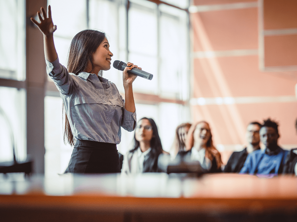 A government official giving a speech on a mic with an audience in the background.