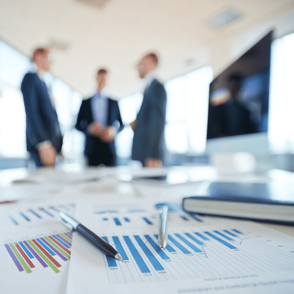 Sheets of graphs and charts on a table with computer and employees in the background.