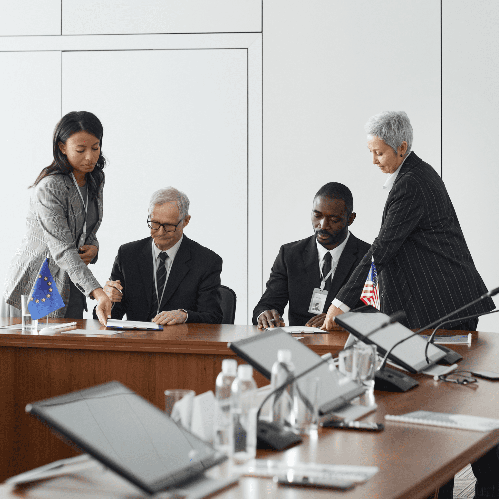 Four government officials at a table signing paperwork while tablets and monitors are shown.