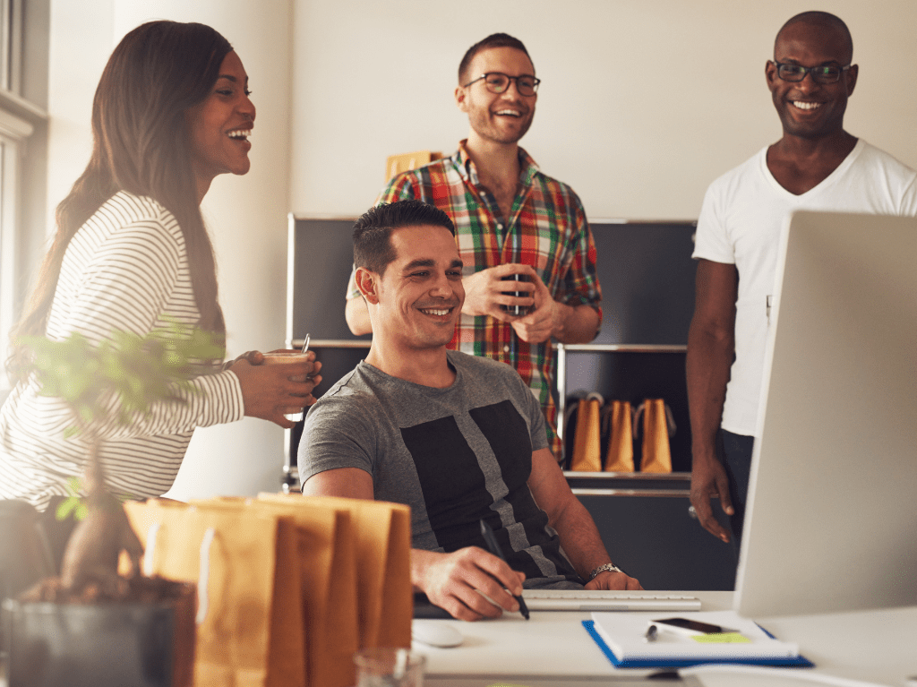 Four associates in an office setting around a computer and all smiling and laughing.