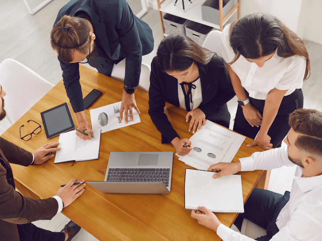 Five professionals sitting around a table working on paperwork with a laptop and tablet.