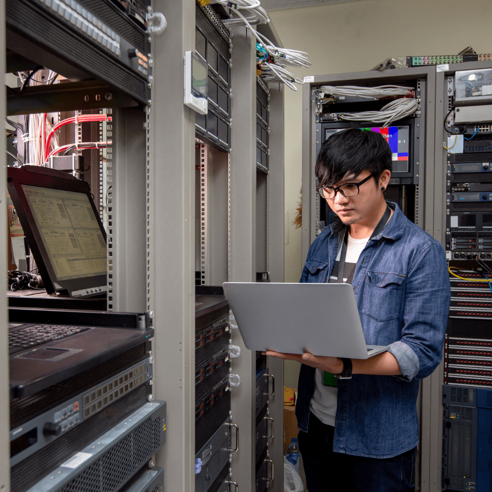 IT specialist working on his laptop in the backroom where multiple servers are housed.